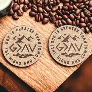 以下が含まれることがあります： 茶色の木目調の丸いコルクコースター2枚。各コースターには、山脈のグラフィックと、「神は高みと低みよりも偉大である」という文字と、「G>A<V」というシンボルが茶色で描かれています。