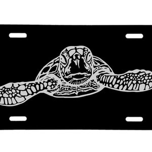 May include: A black and white silhouette of a sea turtle on a black license plate. The turtle is facing the viewer with its head and flippers visible.