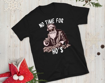 May include: Black t-shirt with a graphic of Santa Claus wearing sunglasses and a suit, holding cash. The text "NO TIME FOR HO'S" is printed above and below the Santa graphic. The shirt is displayed on a wooden surface with holiday decorations.