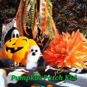 May include: A plush pumpkin with a friendly face, a white ghost, and a black and orange fabric with the text "Pumpkin Patch Kid" are arranged on a black and white fabric with a ghost pattern. The background features a fall scene with orange and green leaves and a bright orange flower.