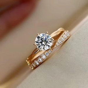 Può includere: Un set di anelli color oro con un anello di fidanzamento solitario e una fede nuziale abbinata. L'anello di fidanzamento ha una grande gemma rotonda e trasparente. La fede è adornata con gemme più piccole e trasparenti. Gli anelli sono su uno sfondo neutro.