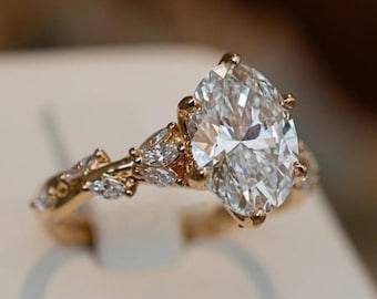 Puede incluir: Un anillo de oro con un gran diamante de talla ovalada como pieza central. La banda presenta delicados detalles en forma de hojas con piedras más pequeñas y brillantes. El anillo se destaca sobre un fondo suave y neutro, resaltando su elegante diseño.