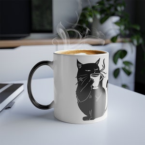 Puede incluir: Una taza de café de cerámica blanca con un asa negra. La taza presenta una ilustración en blanco y negro de un gato sosteniendo una taza de café. El gato tiene una expresión seria en su rostro.