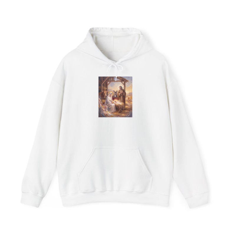 White unisex hoodie with a nativity scene print centered on the chest, depicting Mary, Joseph, and baby Jesus in a stable with warm golden lighting