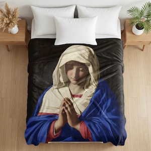 May include: A soft fleece blanket featuring a printed reproduction of a classical painting depicting a figure with a cream-colored head covering, a red garment, and a draped blue cloak, with hands clasped together in a prayerful position.