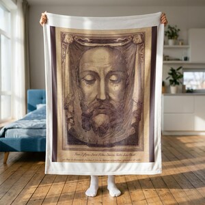 Holy Face plush blanket held up to show its full size in a bright room