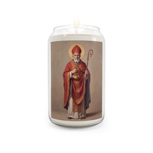 A lit glass jar candle with white wax, featuring a label depicting St. Nicholas dressed in red and gold bishop's robes, holding a golden bag and a pastoral staff, with a golden halo, on a plain white background