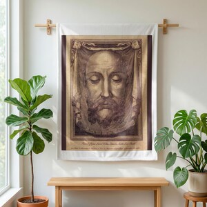 Holy Face plush blanket hung on a wall from a wooden rod above a bench, between potted plants