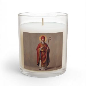 An 11oz glass jar candle with a white wick, featuring a square label depicting St. Nicholas in red and gold bishop's robes, holding a staff and a bag of gold, with a golden halo, on a white background