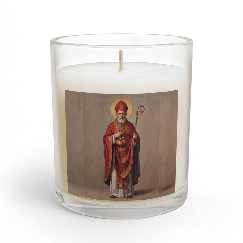 An 11oz glass jar candle with a white wick, featuring a square label depicting St. Nicholas in red and gold bishop's robes, holding a staff and a bag of gold, with a golden halo, on a white background