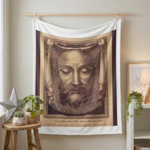 Holy Face plush blanket hung on a wall from a wooden rod above a small plant stand