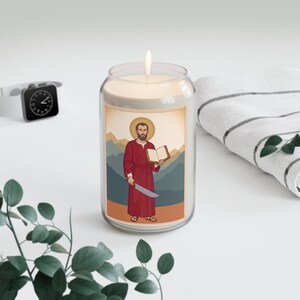 Puede incluir: Una vela en frasco de vidrio transparente con mecha encendida, con una etiqueta que muestra a una figura barbuda con túnica roja sosteniendo un libro abierto y una hoja curva, sobre un fondo de montañas y suelo color canela.