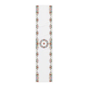 Puede incluir: Un camino de mesa blanco con dos bordes verticales de coloridos patrones florales con elementos rojos, rosas, azules y verdes. En el centro, una corona floral circular rodea un pequeño icono de corazón rojo, creando un diseño simétrico.