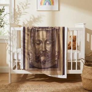 Holy Face plush blanket draped over the side of a white crib in a nursery