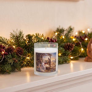 A clear glass candle with a nativity scene label, displayed on a white surface surrounded by evergreen garland, burgundy flowers, pinecones, and warm string lights