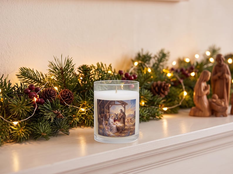 A clear glass candle with a nativity scene label, displayed on a white surface surrounded by evergreen garland, burgundy flowers, pinecones, and warm string lights