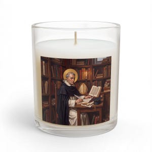 May include: A clear glass candle container filled with white wax, featuring a label depicting a figure in a black habit with a gold halo, writing at a wooden desk in a library filled with books, with a miter and a graduation cap on the desk.