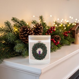 Frosted glass candle with purple candle inside, featuring artwork of a green wreath with red berries, displayed on a white mantelpiece surrounded by evergreen branches, pinecones, and string lights