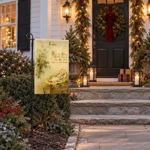 Nativity garden flag displayed on a black stand along a holiday-decorated front entrance, featuring an illustration of baby Jesus in a manger with flowering vines and religious text on a cream-colored fabric