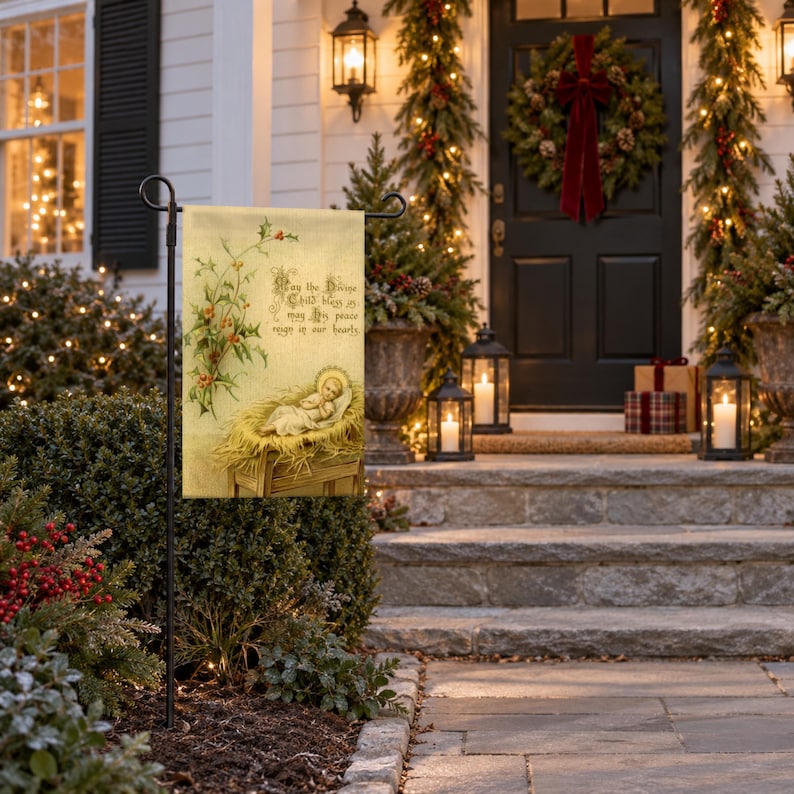 Nativity garden flag displayed on a black stand along a holiday-decorated front entrance, featuring an illustration of baby Jesus in a manger with flowering vines and religious text on a cream-colored fabric