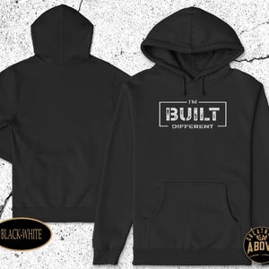 May include: A black hooded sweatshirt featuring a front kangaroo pocket and long sleeves. The chest displays the white text "I'M BUILT DIFFERENT" enclosed within a rectangular border. The back of the hoodie is plain black with no graphic designs.