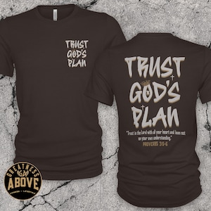 May include: A dark brown short-sleeve t-shirt featuring the text TRUST GOD'S PLAN on the front left chest and a larger version on the back with a crown, two small crosses, and the scripture text Trust in the Lord with all your heart and lean not on your own understanding. PROVERBS 3:5-6.