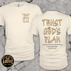 May include: A cream-colored short-sleeve t-shirt featuring the text TRUST GOD'S PLAN on the front left chest. The back displays the same text in larger font with a crown icon, plus the verse Trust in the Lord with all your heart and lean not on your own understanding. PROVERBS 3:5-6.