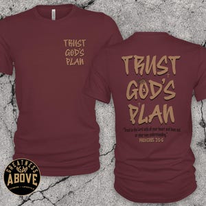 May include: A maroon short-sleeved t-shirt featuring the text TRUST GOD'S PLAN in tan on the left chest and a larger version on the back with the additional text Trust in the Lord with all your heart and lean not on your own understanding. PROVERBS 3:5-6.