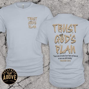May include: A light blue t-shirt featuring the text TRUST GOD'S PLAN in tan on the front left chest. The back displays the same text in a larger font with a crown icon, plus the verse Trust in the Lord with all your heart and lean not on your own understanding. PROVERBS 3:5-6.