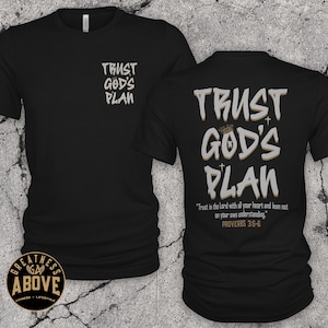May include: A black short-sleeved t-shirt featuring the text "TRUST GOD'S PLAN" in white and tan on the front left chest and a larger version on the back, which also includes the verse "Trust in the Lord with all your heart and lean not on your own understanding. PROVERBS 3:5-6."