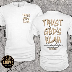 May include: A white short-sleeve t-shirt featuring the text TRUST GOD'S PLAN in tan on the front left chest. The back displays the same text in a larger font with a crown icon, plus the verse Trust in the Lord with all your heart and lean not on your own understanding. PROVERBS 3:5-6.
