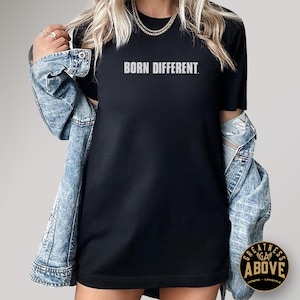 May include: A black short-sleeved t-shirt featuring the text BORN DIFFERENT. printed in a light gray sans-serif font across the chest. The shirt is worn layered under a light blue acid-wash denim jacket with long sleeves and metal buttons.