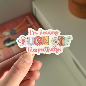 Puede incluir: Una pegatina con el texto "I'm Reading F*CK OFF (Respectfully)" en letras de colores con flores alrededor de las letras. La pegatina está siendo sostenida por una mano.