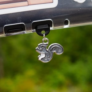 Puede incluir: Un dije plateado con forma de ardilla cuelga de una funda de teléfono. La funda del teléfono tiene pegatinas de una librería, calabazas y un ganso con un suéter. El fondo es follaje verde borroso.