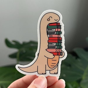 May include: A beige dinosaur sticker with a stack of colorful books. The dinosaur is smiling and holding the books in its arms.