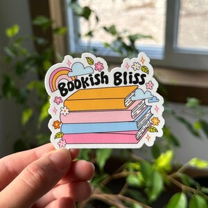 May include: A sticker with a stack of colorful books and the text "Bookish Bliss" in a rainbow arch. The sticker is surrounded by small flowers and stars.