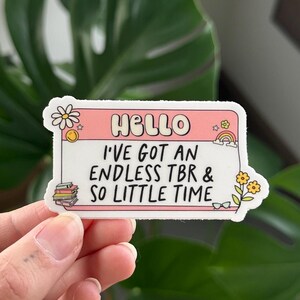 May include: A pink and white sticker with the text "Hello, I've got an endless TBR & so little time" and a stack of books, a daisy, and a rainbow.
