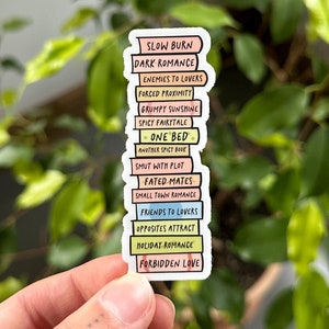 Op de afbeelding: Een sticker met een stapel kleurrijke boeken, elk met een ander romantisch trope erop geschreven. De tropes zijn: Slow Burn, Dark Romance, Enemies to Lovers, Forced Proximity, Grumpy Sunshine, Spicy Fairytale, One Bed, Another Spicy Bove, Smut with Plot, Fated Mates, Small Town Romance, Friends to Lovers, Opposites Attract, Holiday Romance, Forbidden Love.