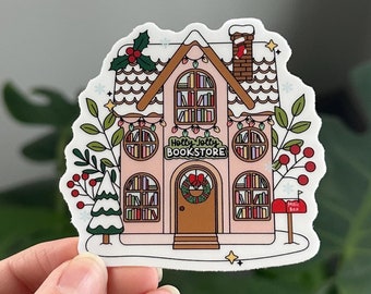 May include: A pink and white sticker of a house with a red door and a wreath. The house has windows with books inside and the text "Holly Jolly Bookstore" above the door. The house is decorated with snow, lights, and holly.