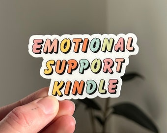May include: A colorful sticker with the words "EMOTIONAL SUPPORT KINDLE" in a retro font. The sticker has a white border and is held by a hand. A small potted plant and a floral curtain are in the background.