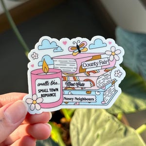 May include: A pink and white sticker with a candle that says "smells like small town romance". The sticker also has a stack of books with the text "County Fair", "Meet Cute", and "Nosey Neighbours".