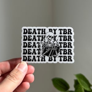 May include: A white sticker featuring black text and a skeleton illustration. The text reads "DEATH BY TBR" repeated five times. The skeleton is depicted reading a book. The sticker is held in a hand.