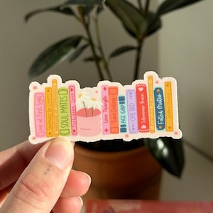 May include: A sticker of a stack of colorful books with titles like "Love at First Sight", "Soul Mates", "Love Triangle", "Fake Dating", "Age Gap", "Overed", "Fated Mates".