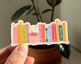May include: A sticker of a stack of colourful books with titles such as "Love at First Sight", "Soul Mates", "Love Triangle", "Fake Dating", "Age Gap", "Overed", "Fated Mates".