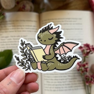 May include: A green dragon with pink wings reading a book. The dragon is sitting on a white background with green leaves and pink flowers. The sticker is on a book.