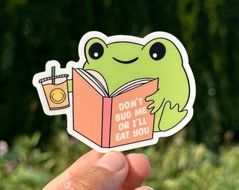 Op de afbeelding: Een groene kikker sticker die een boek en een kopje ijskoffie vasthoudt. Het boek zegt "Stoor me niet, anders eet ik je op".