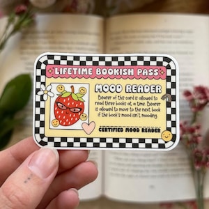 Könnte beinhalten: Ein gelber und schwarzer karierter Aufkleber mit einer pinkfarbenen und roten Erdbeer-Cartoonfigur, die eine Sonnenbrille trägt. Der Aufkleber lautet "LIFETIME BOOKISH PASS" und "MOOD READER". Der Text lautet "Der Inhaber dieser Karte darf drei Bücher gleichzeitig lesen. Der Inhaber darf zum nächsten Buch wechseln, wenn die Stimmung des Buches nicht passt. ZERTIFIZIERTER STIMMUNGSLESER."