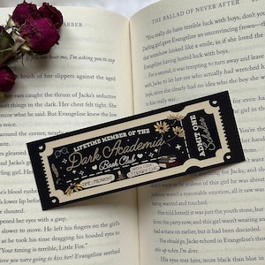 May include: A close-up of an open book with a ticket for a "Dark Academia Bash Club" event. The book is open to a page with the text "The Ballad of Never After" and a scene from the story. The ticket is a black and white design with gold lettering.