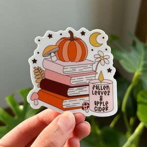 Puede incluir: Una pegatina con una pila de libros, una calabaza, una vela y un hongo. El texto en la pegatina dice "Fallen Leaves & Apple Cider".