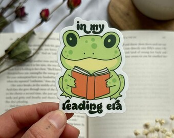 Op de afbeelding: Een groene kikker sticker met een boek in zijn handen. De sticker zegt "in my reading era".